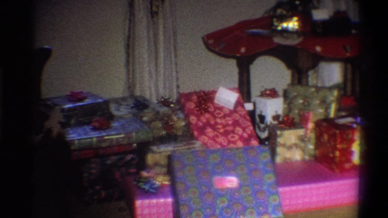 1973 Troy, New York home-movie frame showing colorful wrapped presents arranged beside curtains and a red-topped table.