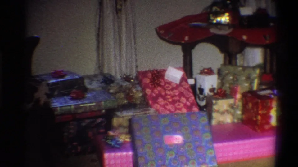 1973 Troy, New York home-movie frame showing colorful wrapped presents arranged beside curtains and a red-topped table.