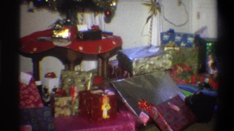 1973 Troy, New York frame showing a gift display with tinsel, wrapped packages, and a small decorated holiday tree at the edge of the room.