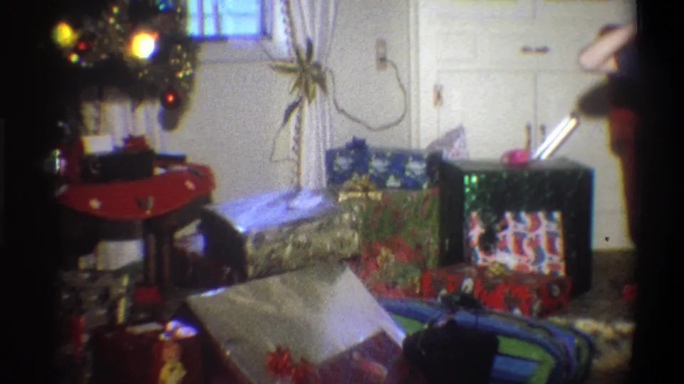 1973 Troy, New York frame of a holiday living room with stacked presents, glowing tree lights, curtains, and a partial figure at the right edge.