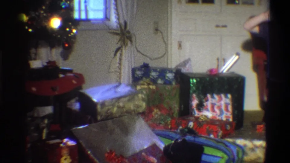 1973 Troy, New York home-movie still showing wrapped Christmas presents, tree lights, patterned curtains, and a person partly cropped at frame right.