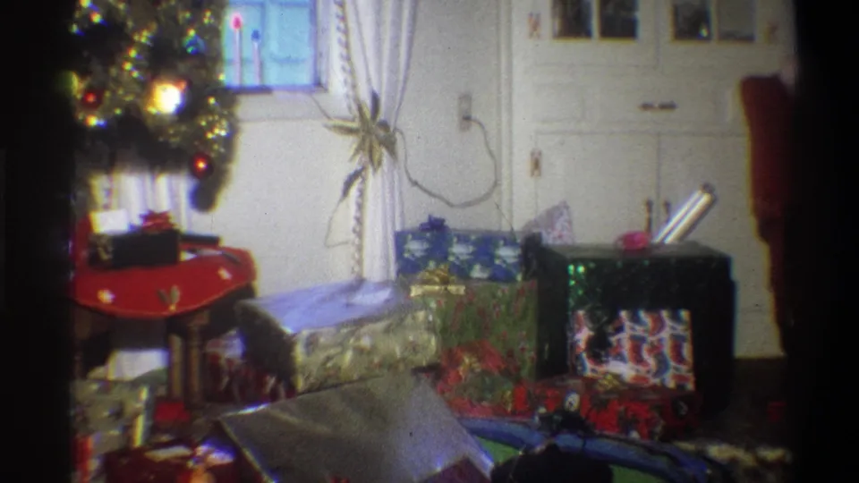 1973 Troy, New York frame with a decorated Christmas tree, many wrapped gifts, curtains, cabinets, and a cropped person at the right edge.