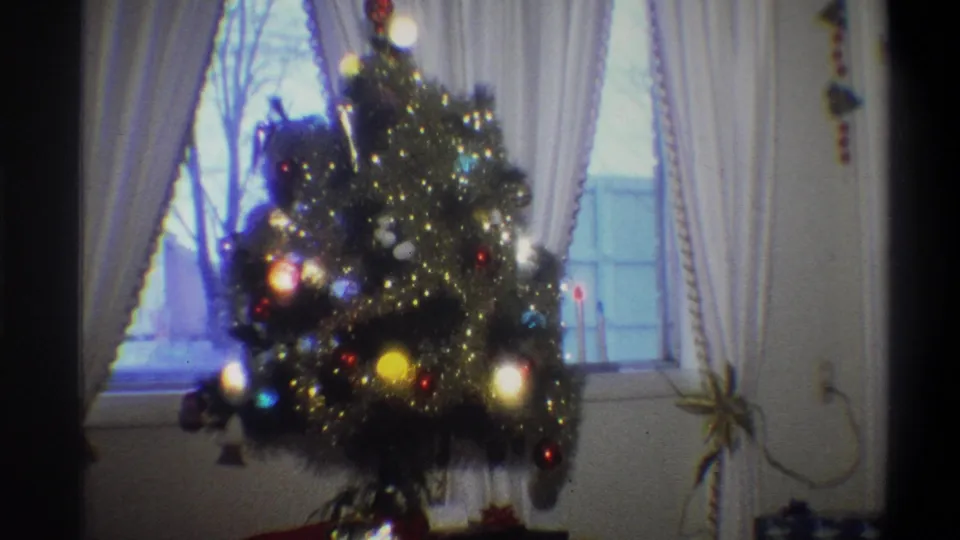 1973 Troy, New York 8mm frame showing a lit Christmas tree with ornaments and tinsel before white curtains and a window.