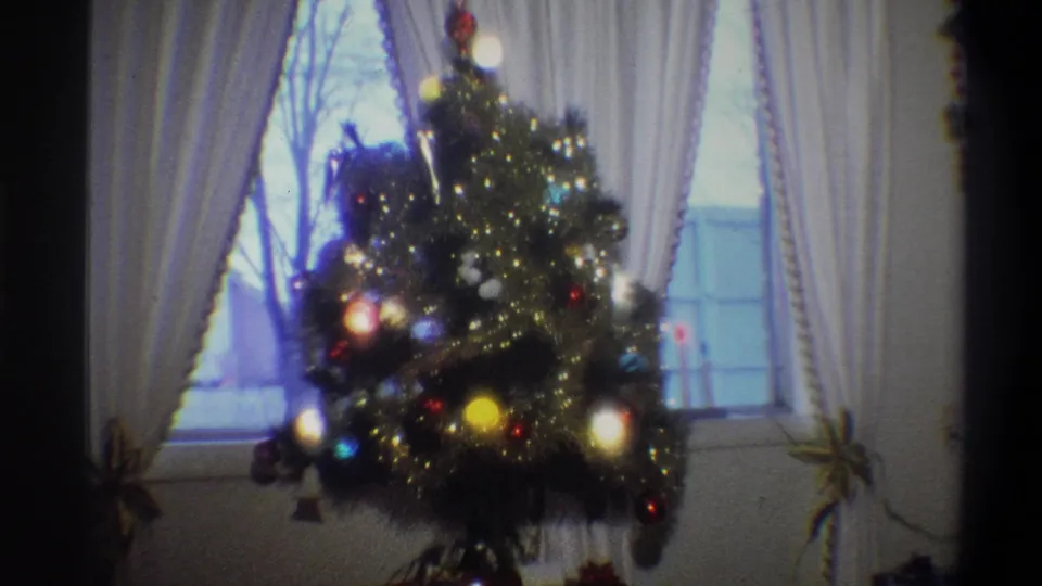 Handheld 8mm view of a decorated Christmas tree glowing beside white curtains in the Troy home interior.