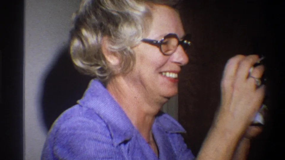 Older woman in a purple collared shirt and round glasses smiles while holding a small camera in Troy, New York, 1973 home footage.