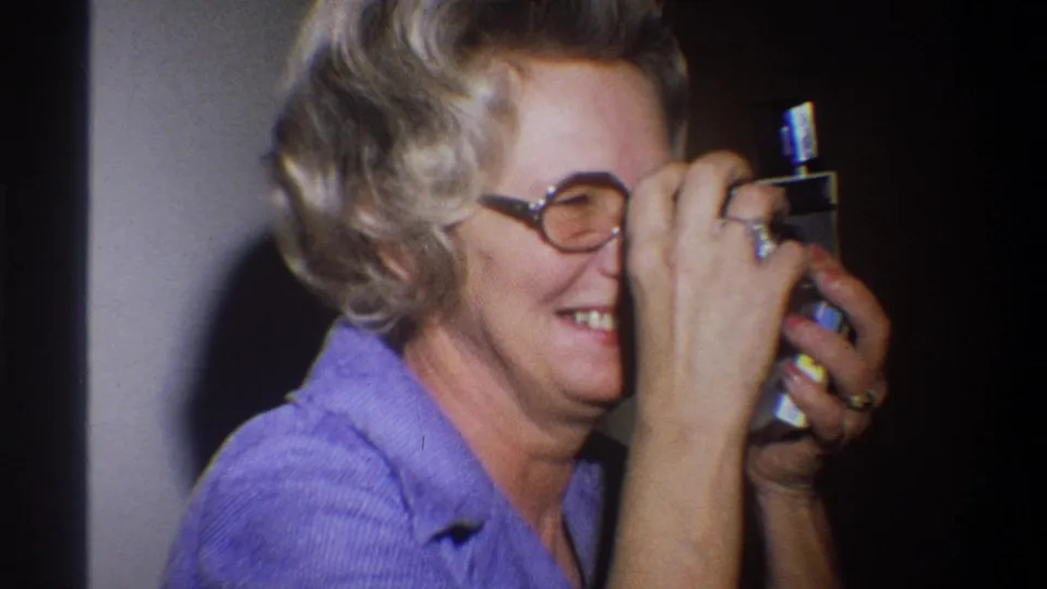 Older woman in round glasses raises a small camera near her face while smiling in 1973 Troy, New York home-movie footage.
