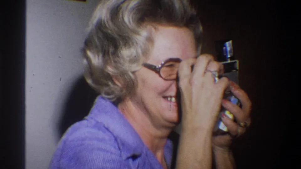 Older woman in a purple shirt looks through a compact camera, smiling in a private Troy, New York interior from 1973 footage.