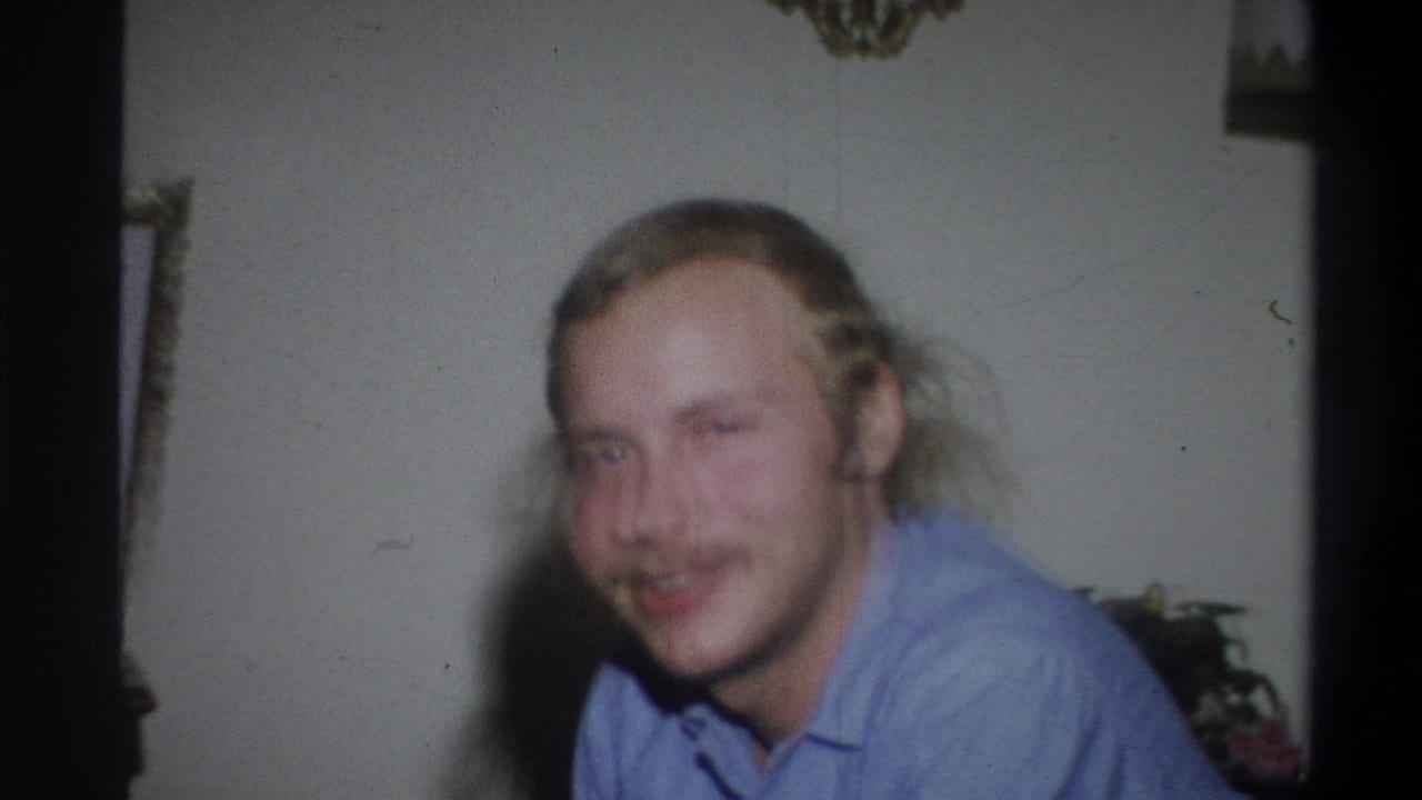 Young man with long blond hair and mustache smiles indoors in a blue collared shirt.