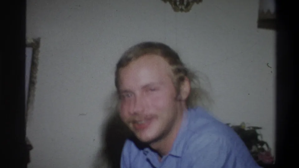 In 1973 Troy, New York home-movie footage, a young man with long blond hair and a mustache faces the camera indoors.
