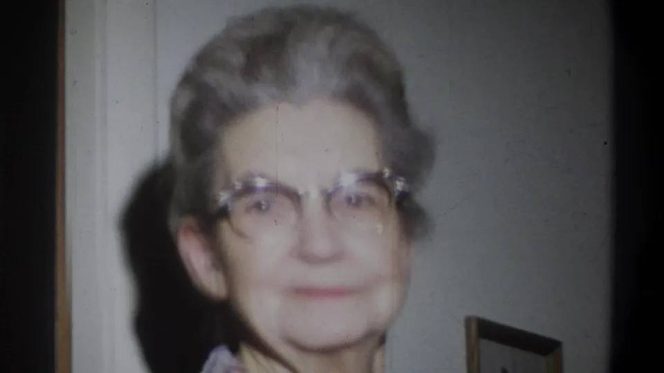An older woman with short gray hair and horn-rimmed glasses looks toward the camera in the Troy, New York home interior.
