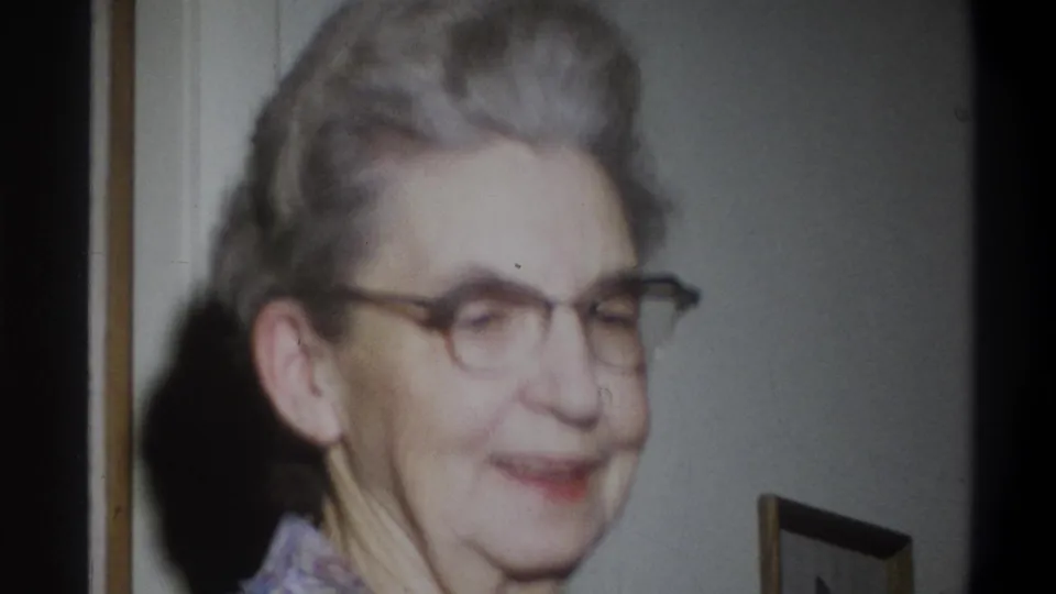 The older woman smiles gently in a side-facing view, with a framed picture partly visible in the private home interior.