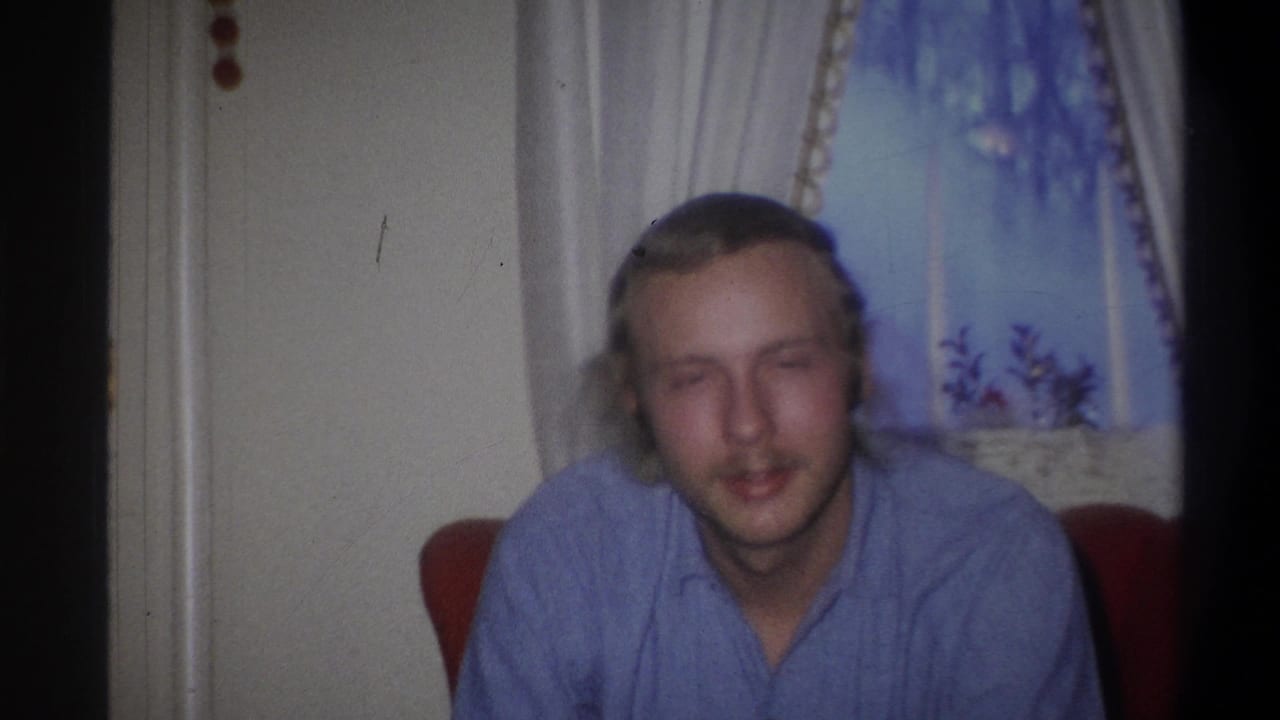 In 1973 Troy, New York home-movie footage, a blond man in a blue shirt sits near white curtains and looks toward the camera.
