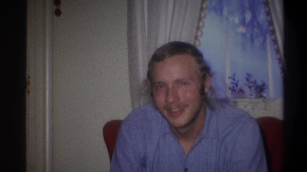 Smiling blond man in a blue button-up shirt seated against a curtained window.
