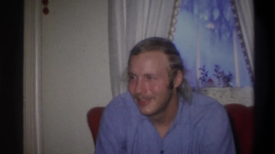 The seated man turns his face to the side, showing his mustache, longer hair, blue shirt, and red chair in the home interior.