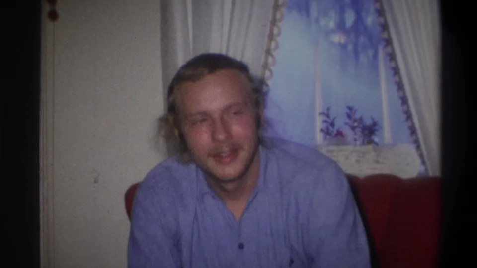 The man faces forward again in the Troy home-movie clip, seated on red furniture with pale curtains and a decorated window behind him.
