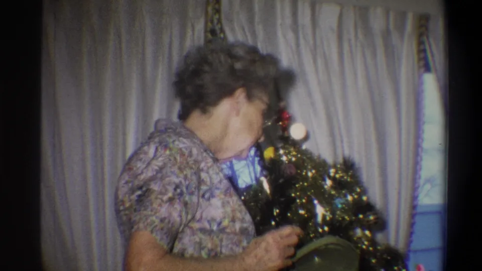 Older woman in a floral dress and glasses holds a round gift box beside a small decorated Christmas tree in a Troy home interior.