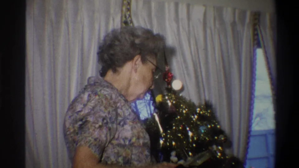 The older woman leans toward the decorated Christmas tree, appearing to inspect or explain the round gift box in the 1973 home-movie scene.