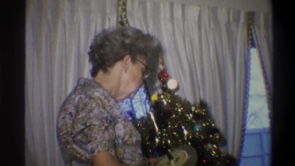 Final frame shows the older woman still beside the small decorated tree, with pale curtains and a window in the private home interior.