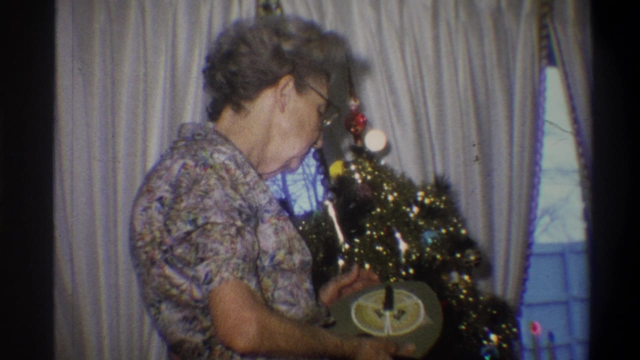 Older woman in glasses adjusts a small Christmas tree beside curtains.