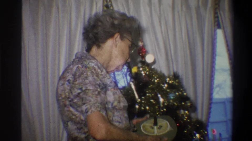 The 1973 Troy, New York frame shows an older woman leaning toward a small artificial Christmas tree in a curtained room.
