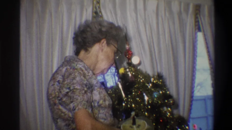A soft 8mm frame shows the woman bending over a small tinsel Christmas tree, with pale curtains and a window behind her.