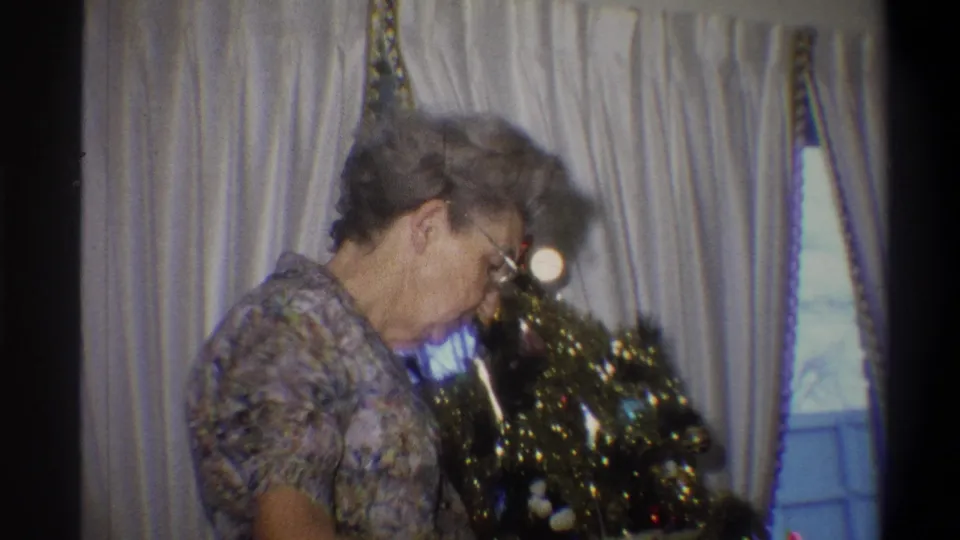 The final frame shows the older woman at the small decorated tree, filmed indoors with grainy 1970s 8mm color texture.