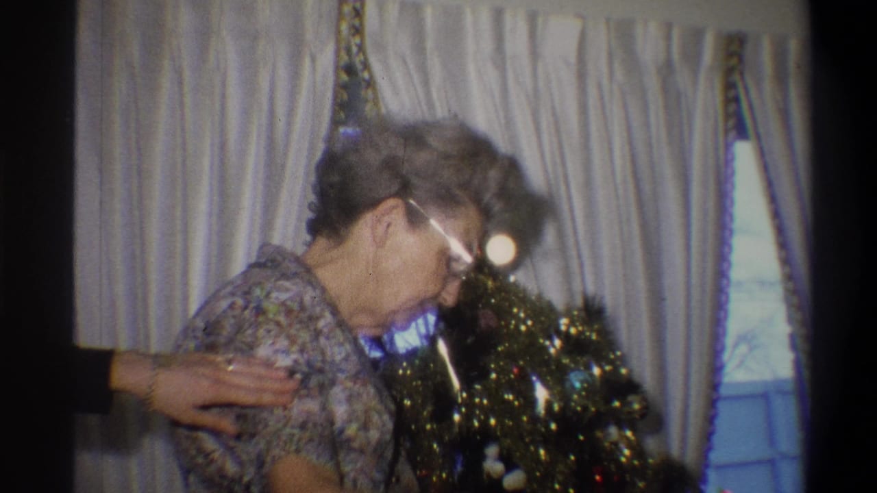 1973 Troy, New York home-movie frame showing an elderly woman beside a small Christmas tree in a private interior.