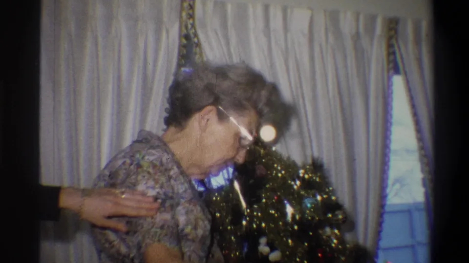 1973 Troy, New York home-movie frame showing an elderly woman beside a small Christmas tree in a private interior.