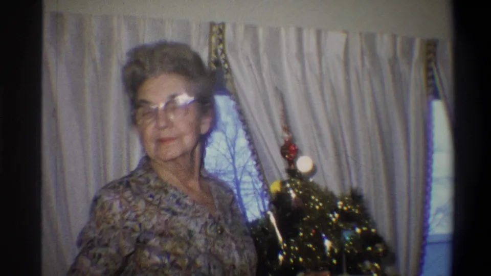 1973 Troy, New York frame of an elderly woman posing beside a small decorated Christmas tree and curtained window.