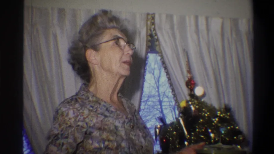 1973 Troy, New York home-movie frame with the woman in profile by a small Christmas tree inside a private room.
