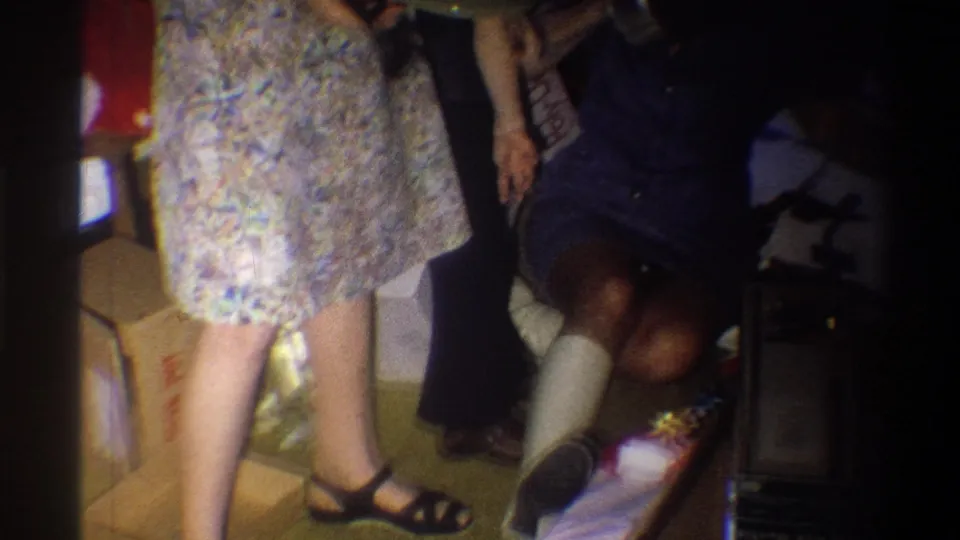 1973 Troy, New York frame with a standing woman in a floral dress beside a seated woman with a leg cast and holiday boxes.