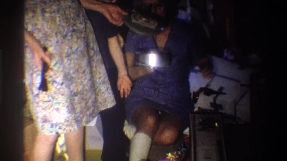 1973 Troy, New York home-movie frame of a seated woman with a leg cast holding a shiny appliance near a woman in a floral dress.