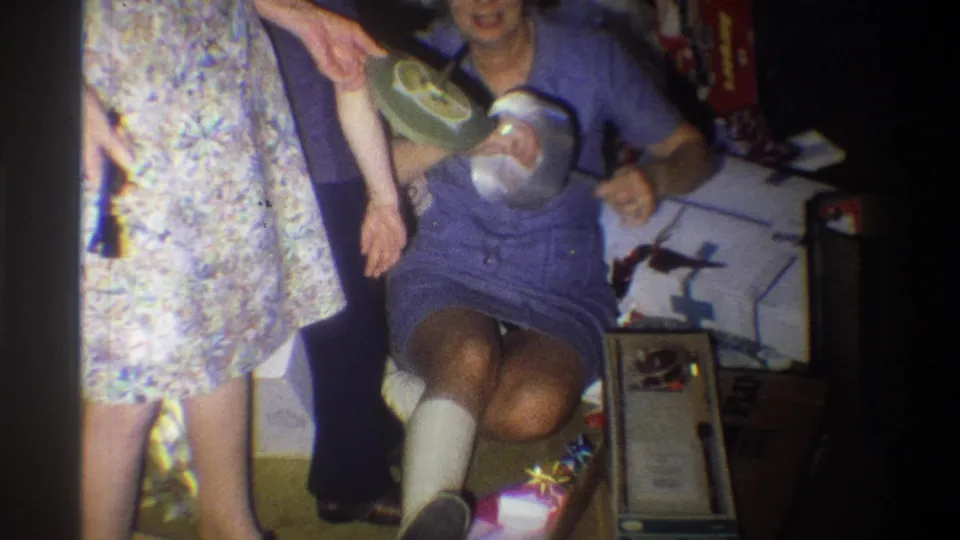 1973 Troy, New York frame showing a smiling seated woman with a leg cast holding a percolator or coffeemaker amid Christmas gifts.