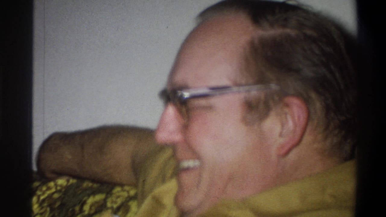 Older man in glasses laughs on a green patterned couch.