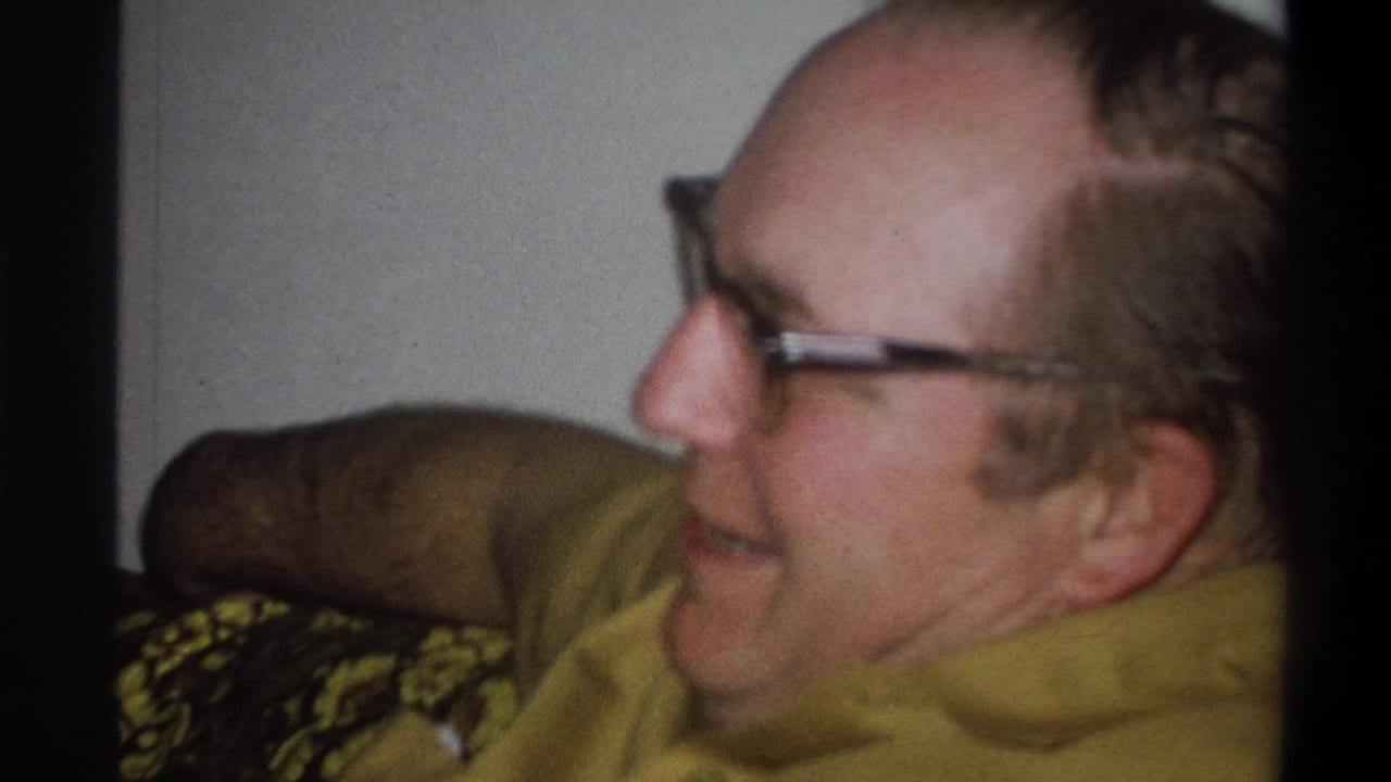 In a Troy, New York home interior, the man sits in profile on the couch with glasses and a yellow shirt visible.