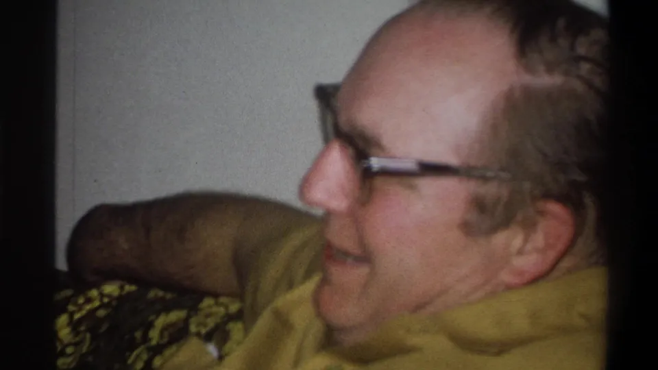 In a Troy, New York home interior, the man sits in profile on the couch with glasses and a yellow shirt visible.