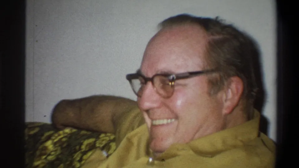 The smiling man leans back on the patterned green couch, his glasses, yellow shirt, and relaxed posture clearly visible.