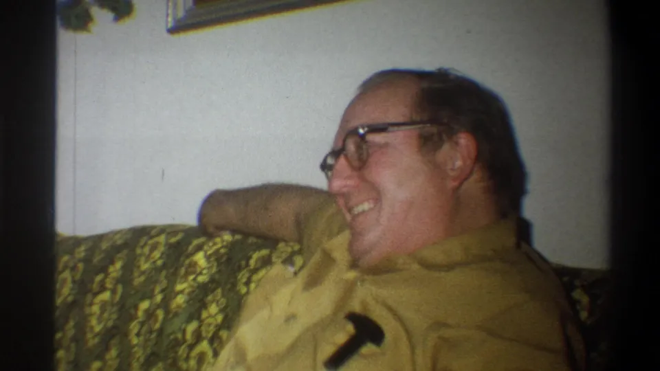 The man laughs while seated on the green couch, with a dark pocket protector visible on his yellow shirt.