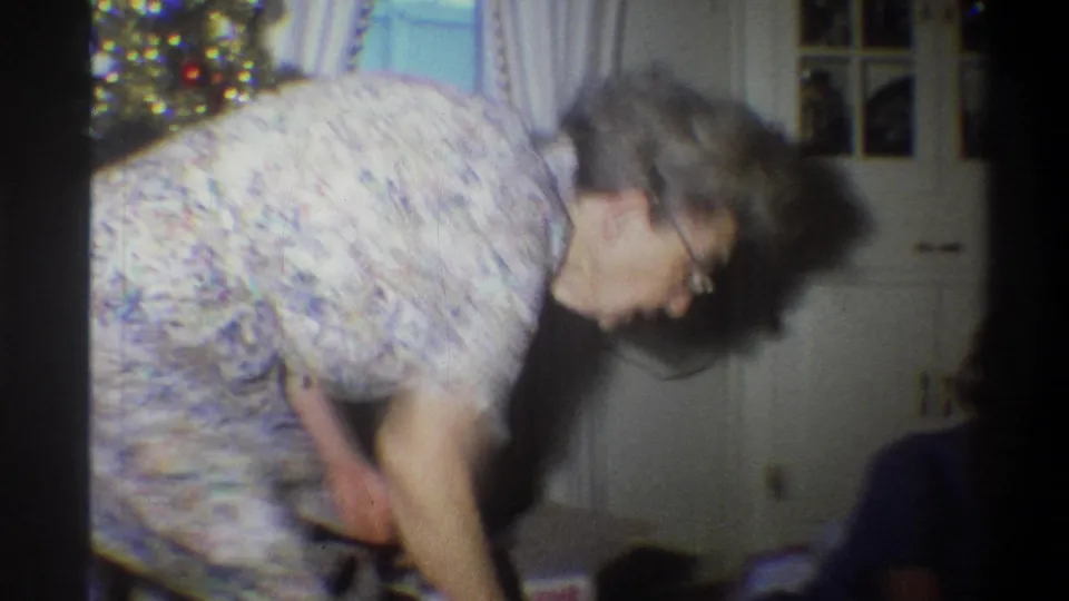 An older woman in a patterned dress bends near a lit Christmas tree inside a Troy, New York home in 1973.