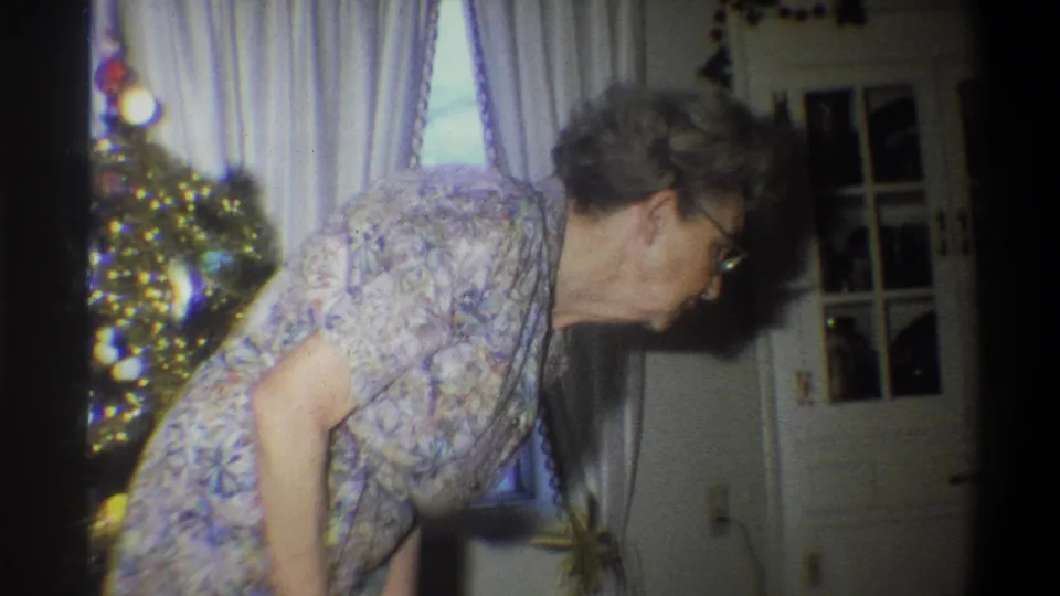 The older woman remains bent forward beside the lit Christmas tree, with curtains and interior doors visible behind her.
