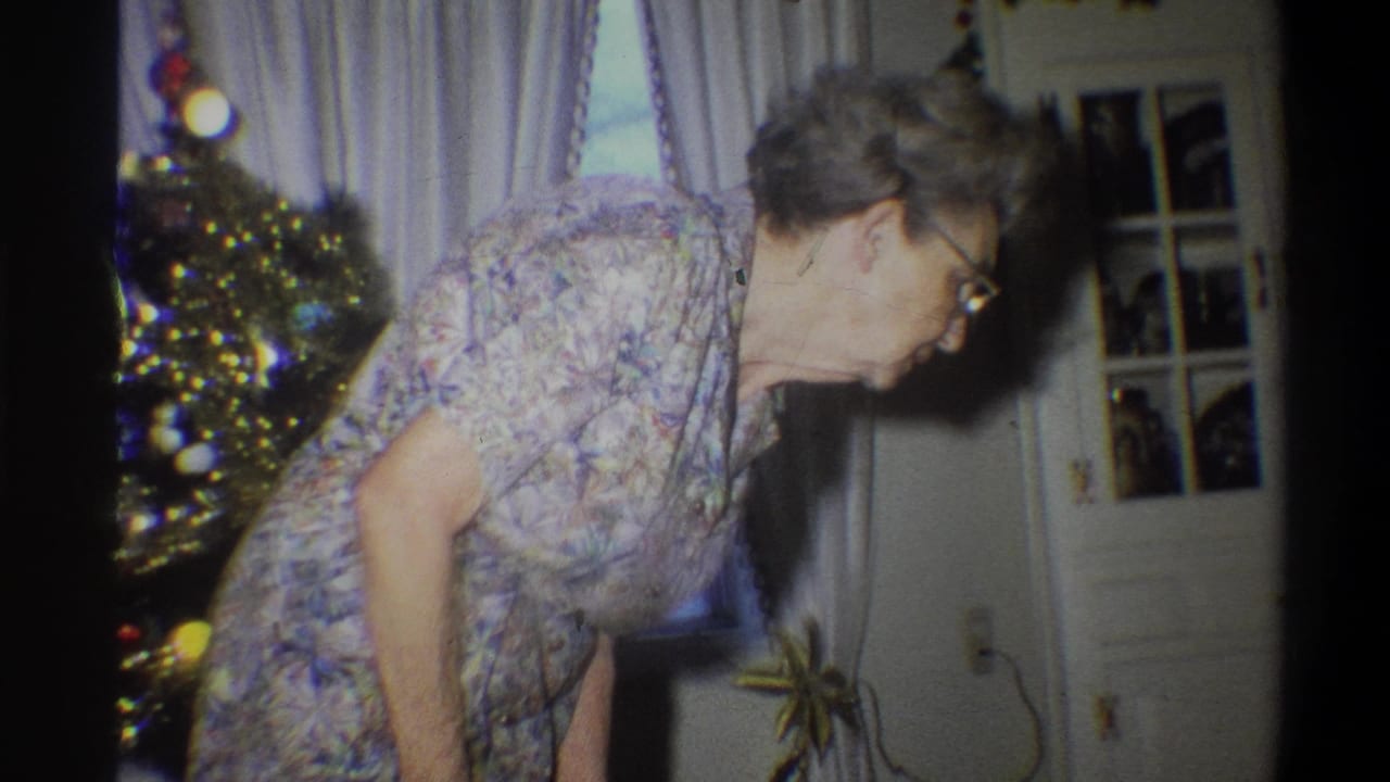 Elderly woman in glasses bends beside a decorated Christmas tree in a living room.