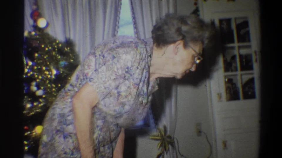 1973 Troy, New York home-movie frame showing an elderly woman in a floral dress bending near a Christmas tree and curtained window.