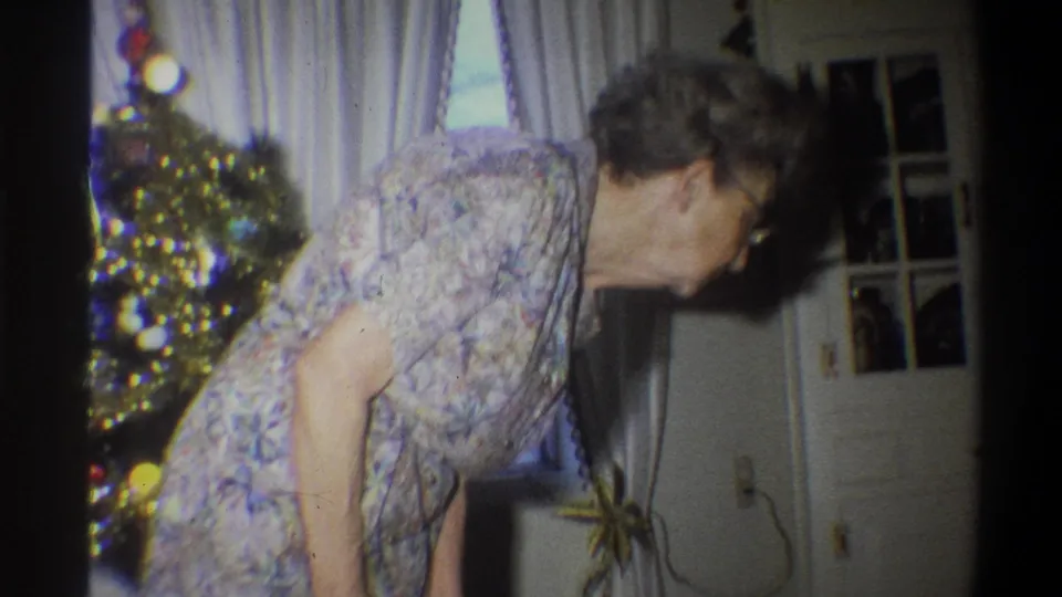 1973 Troy, New York home-movie still showing the woman bending lower beside a Christmas tree and pale curtains.