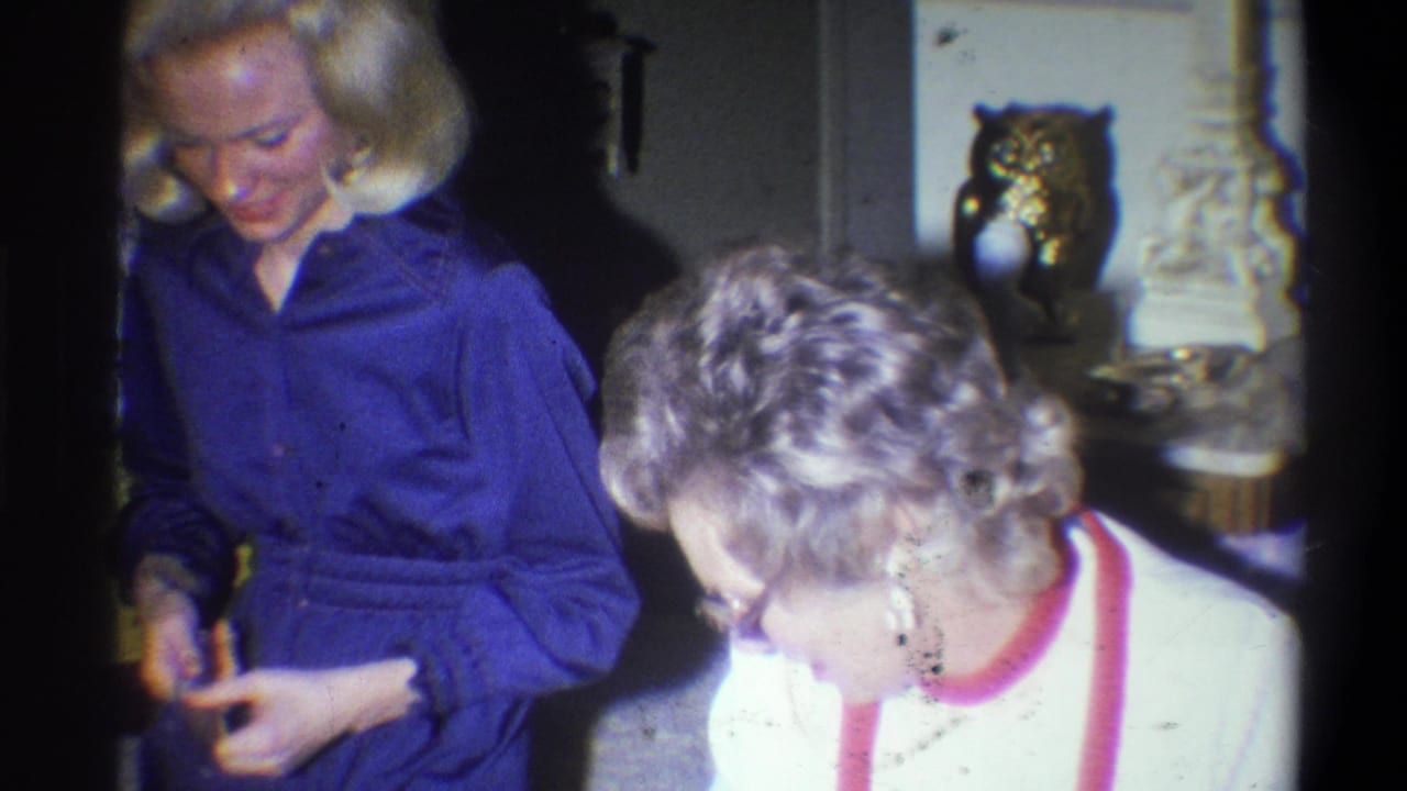 In a 1973 Troy, New York home-movie frame, two adult women stand or sit close together in a living room and look downward.