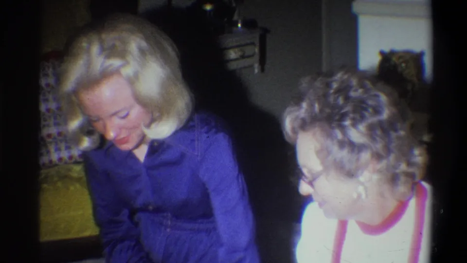 A 1973 home-movie frame shows two adult women seated close in a private Troy living room with patterned upholstery behind them.