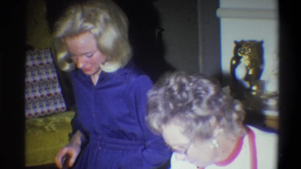 In this 1973 Troy home-movie still, two adult women look downward in a living room with an owl figurine and upholstered furniture nearby.