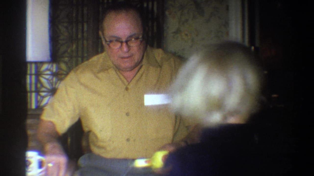 1973 Troy, New York 8mm frame showing a seated man in a tan shirt and glasses with a child partly blocking the foreground.