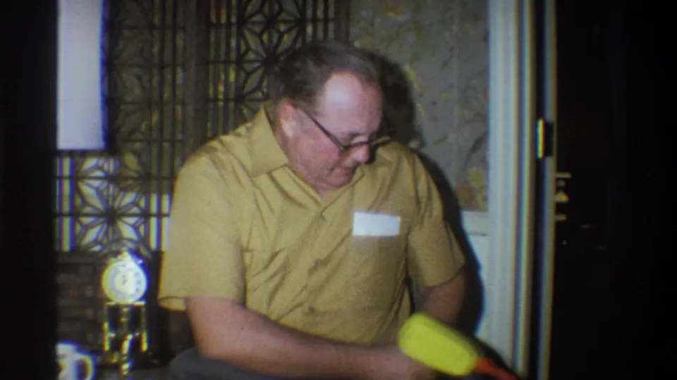 1973 Troy, New York home-movie frame of a man in glasses seated beside a decorative partition, looking toward a yellow toy.