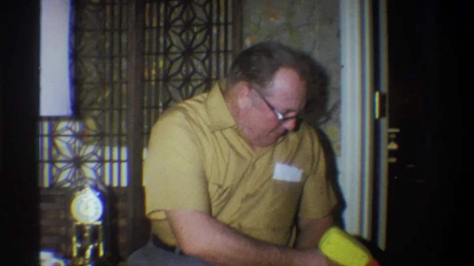 1973 Troy, New York 8mm frame showing a seated man leaning forward near a bright yellow toy, with patterned wall decor behind him.
