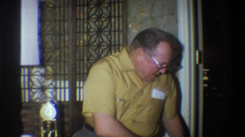 1973 Troy, New York home-movie frame of a man leaning over near a yellow toy, with floral wallpaper and a decorative partition visible.